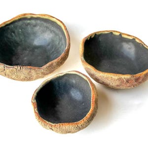 Könnte beinhalten: Drei Keramikschalen mit rustikalem, strukturiertem Äußeren und glattem, dunkelgrauem Inneren. Die Schalen sind unterschiedlich groß und auf einem weißen Hintergrund angeordnet.