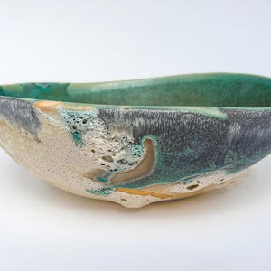 May include: A handcrafted ceramic bowl with an irregular form. The bowl displays a mix of teal, grey, and beige hues, with a textured finish. The inside is a solid teal, and the outside has a marbled appearance. A decorative piece.