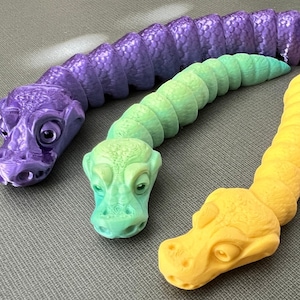 Può includere: Tre serpenti giocattolo colorati, segmentati, con teste dettagliate. I serpenti sono viola, verde e giallo.