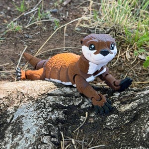 May include: A brown and white otter figurine lying on a log. The toy otter has a detailed design with a brown body, white chest, and a brown head with black eyes. The otter is posed in a relaxed position.
