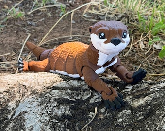 Peut inclure: Figurine de loutre marron et blanche allongée sur une bûche. La loutre jouet a un design détaillé avec un corps marron, une poitrine blanche et une tête marron avec des yeux noirs. La loutre est posée dans une position détendue.