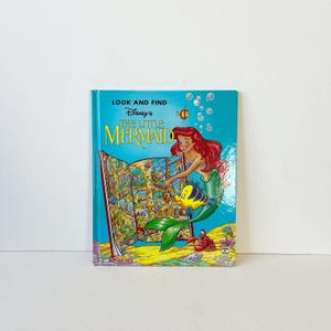 May include: A hardcover "Look and Find" book featuring Disney's The Little Mermaid. The cover is turquoise with Ariel, Flounder, and Sebastian illustrations. The open pages reveal a colorful seek-and-find scene.