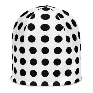 Può includere: Un berretto beanie con una base di colore bianco e un motivo ripetuto di pois neri circolari e pieni. Il berretto presenta una corona arrotondata e un bordo inferiore dritto, mostrando un design grafico coerente su tutta la superficie.