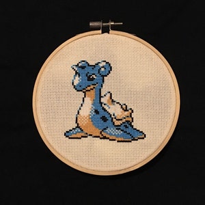 Puede incluir: Una puntada cruzada bordada en azul y amarillo de un personaje de Pokemon, una criatura azul y amarilla similar a un dinosaurio con una concha en la espalda, en un aro de bordado de madera.