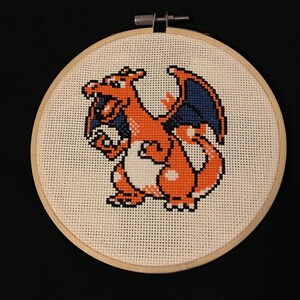 May include: A cross-stitch embroidery hoop featuring a pixelated image of a red and orange dragon with blue wings.
