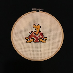 May include: A cross-stitch embroidery hoop with a yellow and red pixelated turtle design.