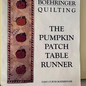 May include: A printed pattern for a pumpkin patch table runner. The runner features three pumpkin appliqués in shades of orange and red on a patterned background. The text reads "Curtis Boehringer Quilting" and "The Pumpkin Patch Table Runner."