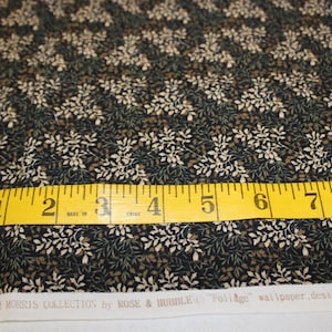 May include: A close-up of black fabric with a white and brown floral pattern. A yellow measuring tape is placed on top of the fabric, showing measurements from 1 to 9 inches. The text "THE WILLIAM MORRIS COLLECTION by ROSE & HUBBLE "Foliage" wallpaper, designed by John Henry Dearle" is visible at the bottom of the image.
