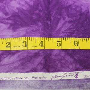May include: Purple fabric with a tie-dye pattern. A measuring tape shows the fabric is 8 inches wide. The fabric has a white label with the text "Color Connectors by Heide Stoll-Weber for Free Spirit Studio Collection".