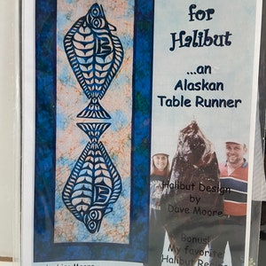 May include: An Alaskan table runner with a blue and white fish design. The text "Hungry for Halibut" is displayed above the fish design. The table runner is designed by Lisa Moore of Quilts With a Twist.