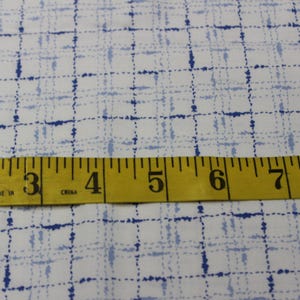 May include: White fabric with a blue and light blue checkered pattern. A yellow measuring tape with the numbers 2 through 8 is placed across the fabric.