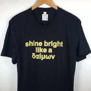 May include: Black t-shirt with gold lettering that reads "shine bright like a &delta;&alpha;ί&mu;&omega;&nu;".