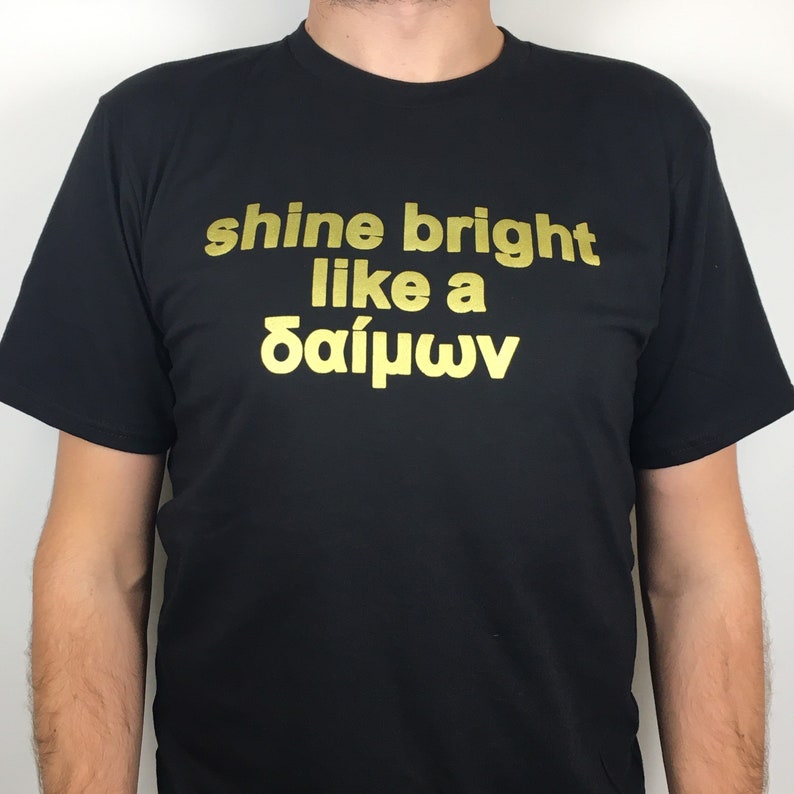 May include: Black t-shirt with gold lettering that reads "shine bright like a &delta;&alpha;ί&mu;&omega;&nu;".