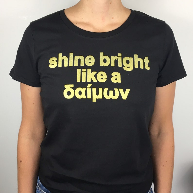 May include: Black t-shirt with gold lettering that reads "shine bright like a &delta;&alpha;ί&mu;&omega;&nu;".