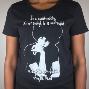 May include: Black t-shirt with a white graphic of Angela Davis, a quote from her that reads "In a racist-society, it's not enough to be non-racist, we must be anti-racist. -Angela Davis"
