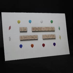 May include: A white birthday card with the Irish Gaelic phrase "Lá Breithe Shona Duit" spelled out in wooden letter tiles. The card is decorated with colorful balloon and confetti illustrations.