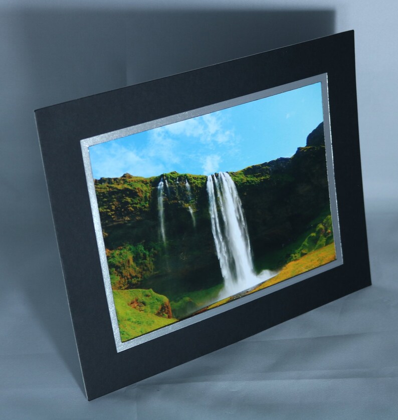 K&ouml;nnte beinhalten: Ein gerahmtes Foto eines Wasserfalls, der eine gr&uuml;ne Klippe hinunterst&uuml;rzt. Der Himmel ist blau mit wei&szlig;en Wolken. Der Rahmen ist schwarz mit einem silbernen Rand.