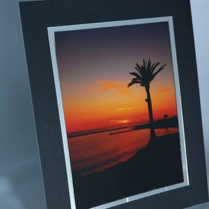 May include: A black picture frame with a silver border, showcasing a photograph of a palm tree silhouetted against a vibrant orange and red sunset over a body of water.