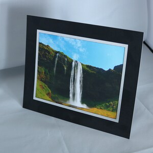 K&ouml;nnte beinhalten: Ein schwarz gerahmtes Foto eines Wasserfalls, der eine gr&uuml;ne Klippe hinunterst&uuml;rzt. Der Himmel ist blau mit wei&szlig;en Wolken.