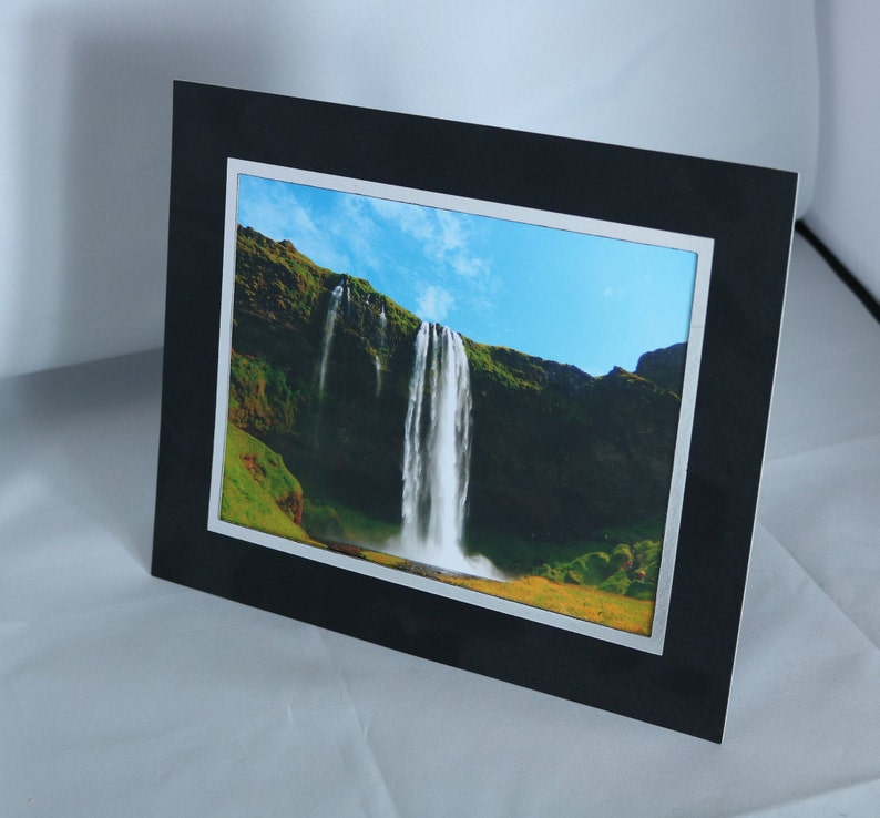 K&ouml;nnte beinhalten: Ein schwarz gerahmtes Foto eines Wasserfalls, der eine gr&uuml;ne Klippe hinunterst&uuml;rzt. Der Himmel ist blau mit wei&szlig;en Wolken.