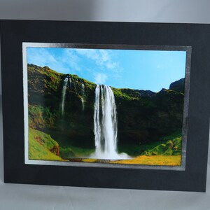 K&ouml;nnte beinhalten: Ein gerahmtes Foto eines Wasserfalls, der eine felsige Klippe hinunterst&uuml;rzt. Das Bild zeigt einen strahlend blauen Himmel mit wei&szlig;en Wolken und &uuml;ppige gr&uuml;ne Vegetation.