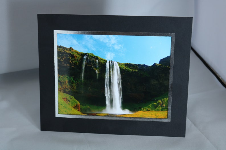 K&ouml;nnte beinhalten: Ein gerahmtes Foto eines Wasserfalls, der eine felsige Klippe hinunterst&uuml;rzt. Das Bild zeigt einen strahlend blauen Himmel mit wei&szlig;en Wolken und &uuml;ppige gr&uuml;ne Vegetation.