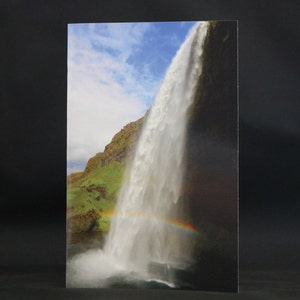 Könnte beinhalten: Ein großer Wasserfall stürzt von einer felsigen Klippe herab, mit einem Regenbogen, der sich über das Wasser wölbt. Der Himmel ist blau mit weißen Wolken.