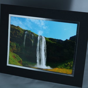 K&ouml;nnte beinhalten: Ein gerahmtes Foto eines Wasserfalls, der eine gr&uuml;ne Klippe hinunterst&uuml;rzt. Der Himmel ist blau mit wei&szlig;en Wolken.