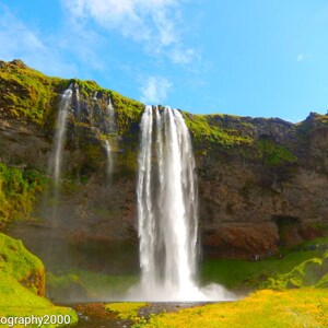 K&ouml;nnte beinhalten: Ein gro&szlig;er Wasserfall st&uuml;rzt sich &uuml;ber eine mit Moos bedeckte Felswand. Das Wasser ist wei&szlig; und sch&auml;umend, und die Klippe ist eine Mischung aus Braun und Gr&uuml;n. Der Himmel ist blau und in der Ferne sind einige Wolken zu sehen.