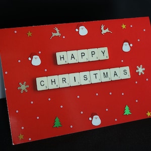 May include: A red card with white dots and a white border. The card says "Happy Christmas" in white letters made from Scrabble tiles. There are also small paper cutouts of Santa, reindeer, stars, and Christmas trees.