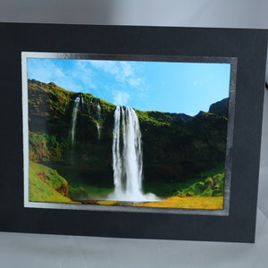 K&ouml;nnte beinhalten: Ein gerahmtes Foto eines Wasserfalls, der eine gr&uuml;ne Klippe hinunterst&uuml;rzt. Der Himmel ist blau mit wei&szlig;en Wolken. Der Rahmen ist schwarz mit einem silbernen Rand.