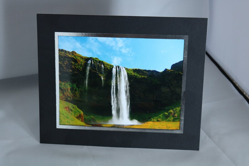 K&ouml;nnte beinhalten: Ein gerahmtes Foto eines Wasserfalls, der eine gr&uuml;ne Klippe hinunterst&uuml;rzt. Der Himmel ist blau mit wei&szlig;en Wolken. Der Rahmen ist schwarz mit einem silbernen Rand.
