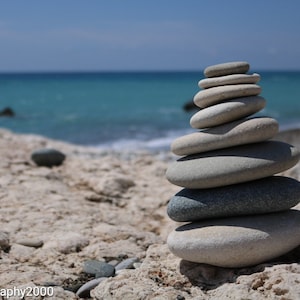 Könnte beinhalten: Ein Stapel glatter, grauer und weißer Steine, die auf einem Sandstrand übereinander balanciert sind. Die Steine sind in Pyramidenform angeordnet, wobei der größte Stein unten und der kleinste Stein oben liegt. Der Hintergrund ist ein blaues Meer und ein blauer Himmel.