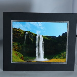 K&ouml;nnte beinhalten: Ein gerahmtes Foto eines Wasserfalls, der eine gr&uuml;ne Klippe hinunterst&uuml;rzt. Der Himmel ist blau mit wei&szlig;en Wolken. Der Rahmen ist schwarz mit einem silbernen Rand.