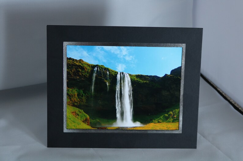 K&ouml;nnte beinhalten: Ein gerahmtes Foto eines Wasserfalls, der eine gr&uuml;ne Klippe hinunterst&uuml;rzt. Der Himmel ist blau mit wei&szlig;en Wolken. Der Rahmen ist schwarz mit einem silbernen Rand.
