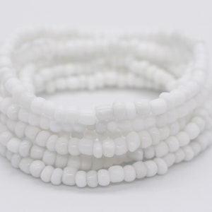 Puede incluir: Un brazalete de cuentas blancas hecho de pequeñas cuentas redondas. El brazalete está envuelto alrededor de sí mismo varias veces, creando un aspecto grueso y en capas.