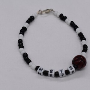 Puede incluir: Pulsera de cuentas negras y blancas con un dije de baloncesto rojo y la palabra "NETS" escrita en cuentas blancas.