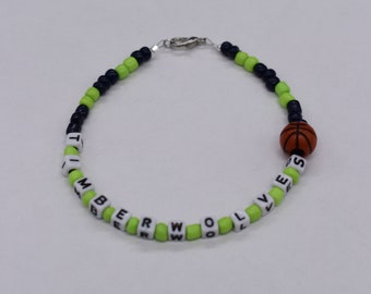 May include: A beaded bracelet featuring the text "TIMBERWOLVES" in white letter beads, interspersed with green and navy beads. A small basketball bead is also included. The bracelet is secured with a silver clasp.