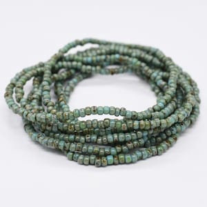 Puede incluir: Un brazalete de cuentas azul turquesa con un acabado moteado. El brazalete está hecho de cuentas pequeñas y redondas y está envuelto alrededor de sí mismo varias veces.