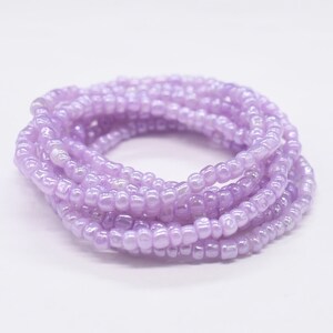 Könnte beinhalten: Eine Spirale aus hellvioletten Rocailles-Perlen. Die kleinen, runden Perlen haben eine leicht schimmernde Oberfläche. Diese Perlen werden wahrscheinlich für Schmuck oder andere Bastelprojekte verwendet.