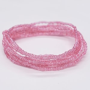 May include: A collection of delicate, light pink seed bead strands, arranged in a circular pattern. The beads are small and evenly sized, creating a smooth, textured appearance. The background is plain white.