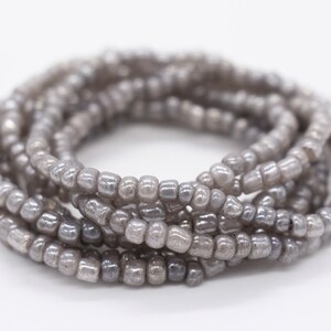 May include: A close-up of a gray beaded bracelet. The bracelet is made of small, round beads that are strung together in a continuous loop. The beads are a light gray color and have a slightly iridescent finish.