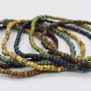 Può includere: Una collezione di sei braccialetti sottili realizzati con piccole perline rotonde multicolori. Le perline presentano una finitura testurizzata e maculata in varie tonalità terrose, tra cui rosso scuro, beige, verde acqua, blu navy, verde oliva e marrone.