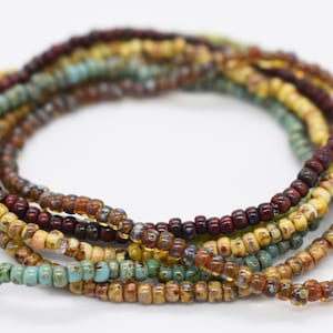 Puede incluir: Varias hebras de pequeñas cuentas de semillas de colores en tonos turquesa, marrón, amarillo y burdeos. Las cuentas están ensartadas, formando collares o pulseras enrolladas. Las cuentas tienen un aspecto ligeramente texturizado.