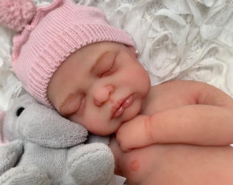 Puede incluir: Una muñeca bebé realista con los ojos cerrados, que lleva un gorro de punto rosa con un pompón. La muñeca sostiene un juguete de elefante de peluche gris. La muñeca está acostada sobre una superficie blanca texturizada.