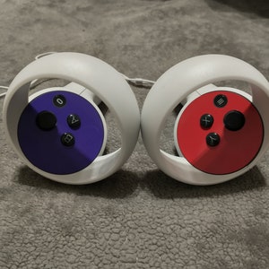 Puede incluir: Dos mandos VR blancos con placas frontales circulares. Un mando tiene una placa frontal morada con botones negros etiquetados como 'A', 'B' y 'X'. El otro mando tiene una placa frontal roja con botones negros etiquetados como 'X', 'Y' y 'A'.