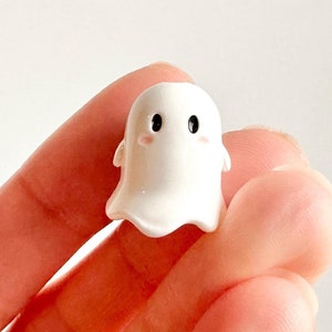 May include: A small white ceramic ghost figurine with black eyes and pink cheeks. The ghost has a rounded head and a flowing, translucent body.