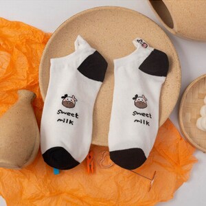 Pu&ograve; includere: Un paio di calzini alla caviglia bianchi con tallone e punta neri. Ogni calzino presenta una grafica di mucca dei cartoni animati e le parole "Sweet milk" in nero. I calzini sono esposti su un piatto beige con carta velina arancione.