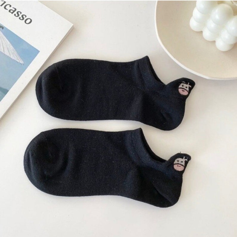 Pu&ograve; includere: Un paio di calzini neri invisibili con un piccolo volto di mucca ricamato sul tallone. I calzini sono realizzati in materiale morbido e sono progettati per essere indossati con le scarpe. Sono appoggiati su una superficie bianca.