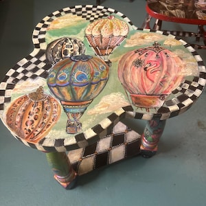 Könnte beinhalten: Ein skurriler, handbemalter Beistelltisch mit einem kleeblattförmigen Oberteil. Der Tisch zeigt bunte Heißluftballons, einen schwarz-weißen Schachbrettmuster-Rand und dekorative Beine. Der Tisch hat einen Durchmesser von ca. 61 cm und ist 46 cm hoch.